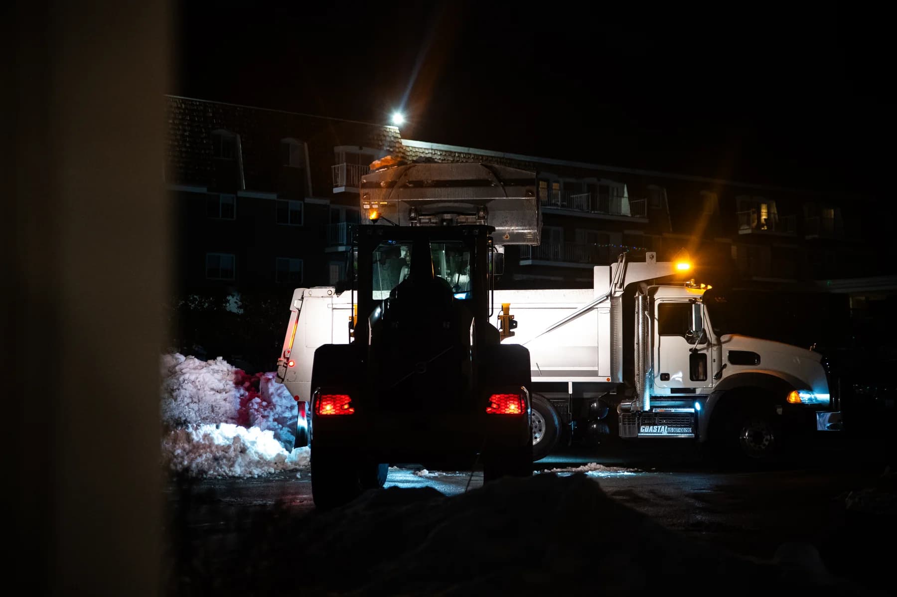 A Coastal Corp wheel loader loads snow into a Mack dump truck at night outside a residential complex.