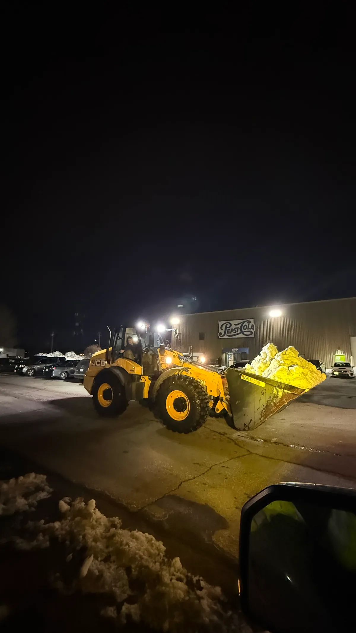 A Coastal Corp wheel loader carries a full bucket of snow past a warehouse during nighttime clearing.