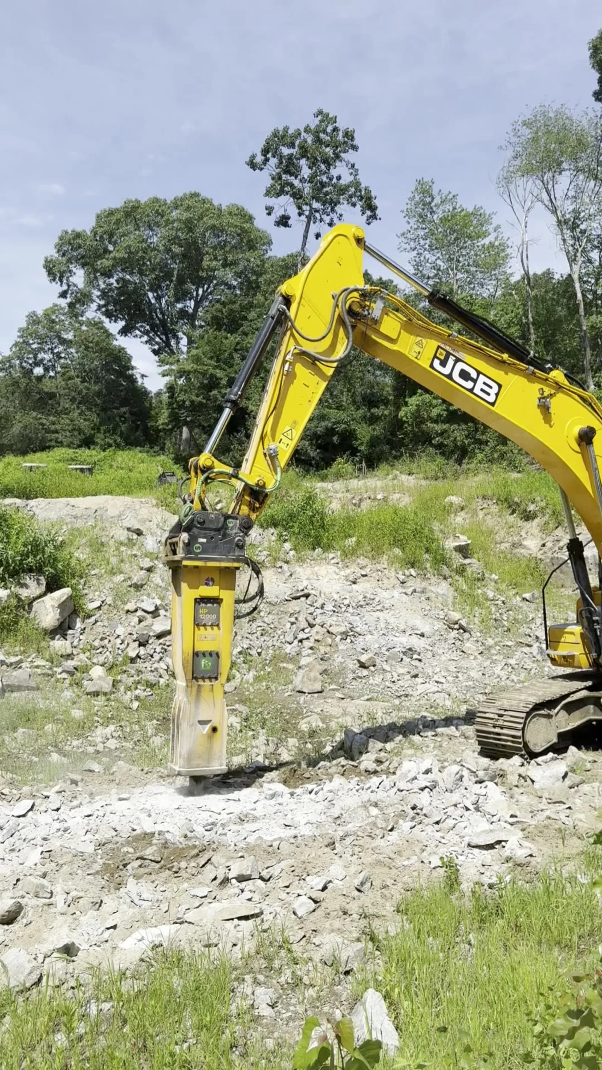 A Coastal Corp JCB tracked excavator runs a 12,000 lb hydraulic breaker, hammering and fracturing ledge rock on a wooded excavation site.
