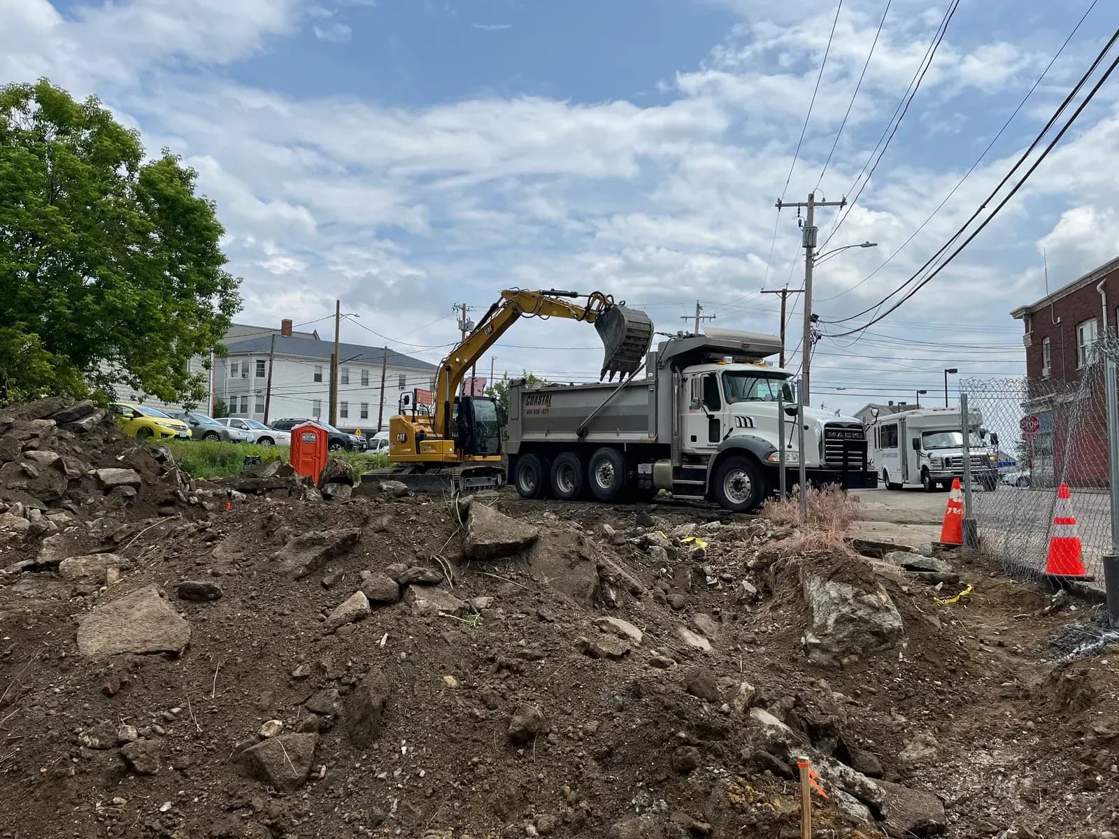 A CAT excavator loads soil into a Coastal Corp Mack tri-axle dump truck at an urban excavation site.