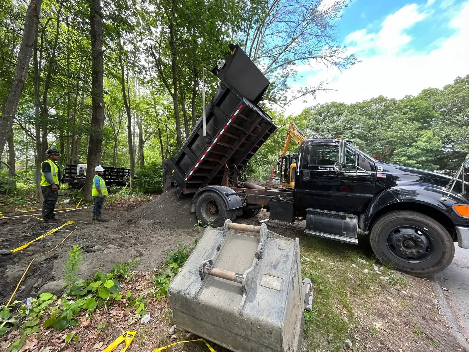 A Coastal Corp dump truck places a load of fill at a wooded jobsite while crew members direct the work and an excavator digs in the background.