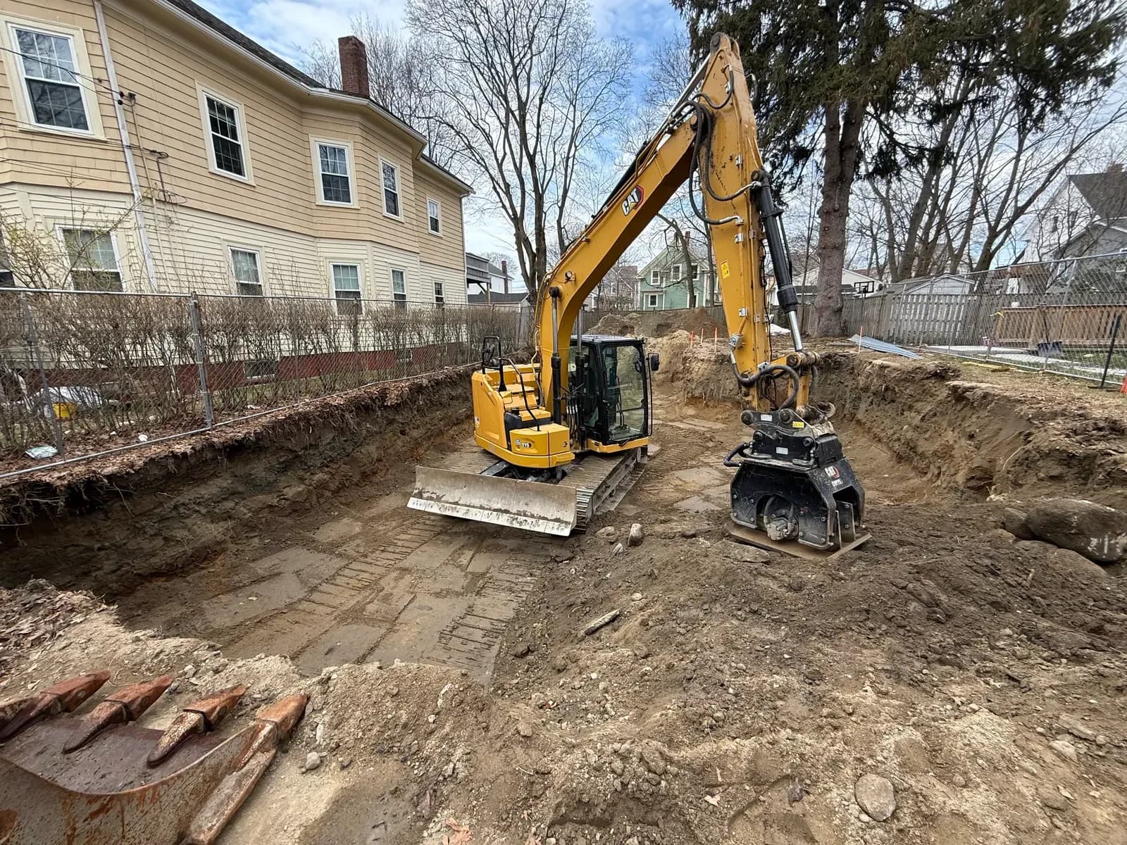 A CAT excavator fitted with a plate compactor works in a deep foundation excavation on a fenced residential lot.