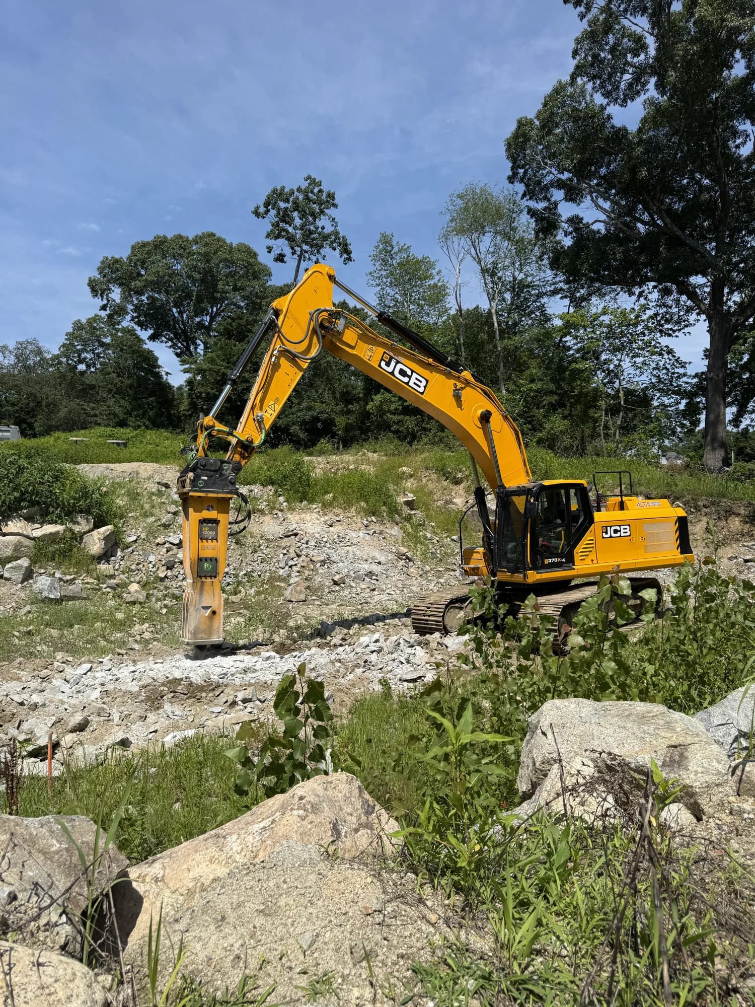 A Coastal Corp JCB tracked excavator with a hydraulic breaker works a rocky excavation surrounded by broken stone and large boulders.