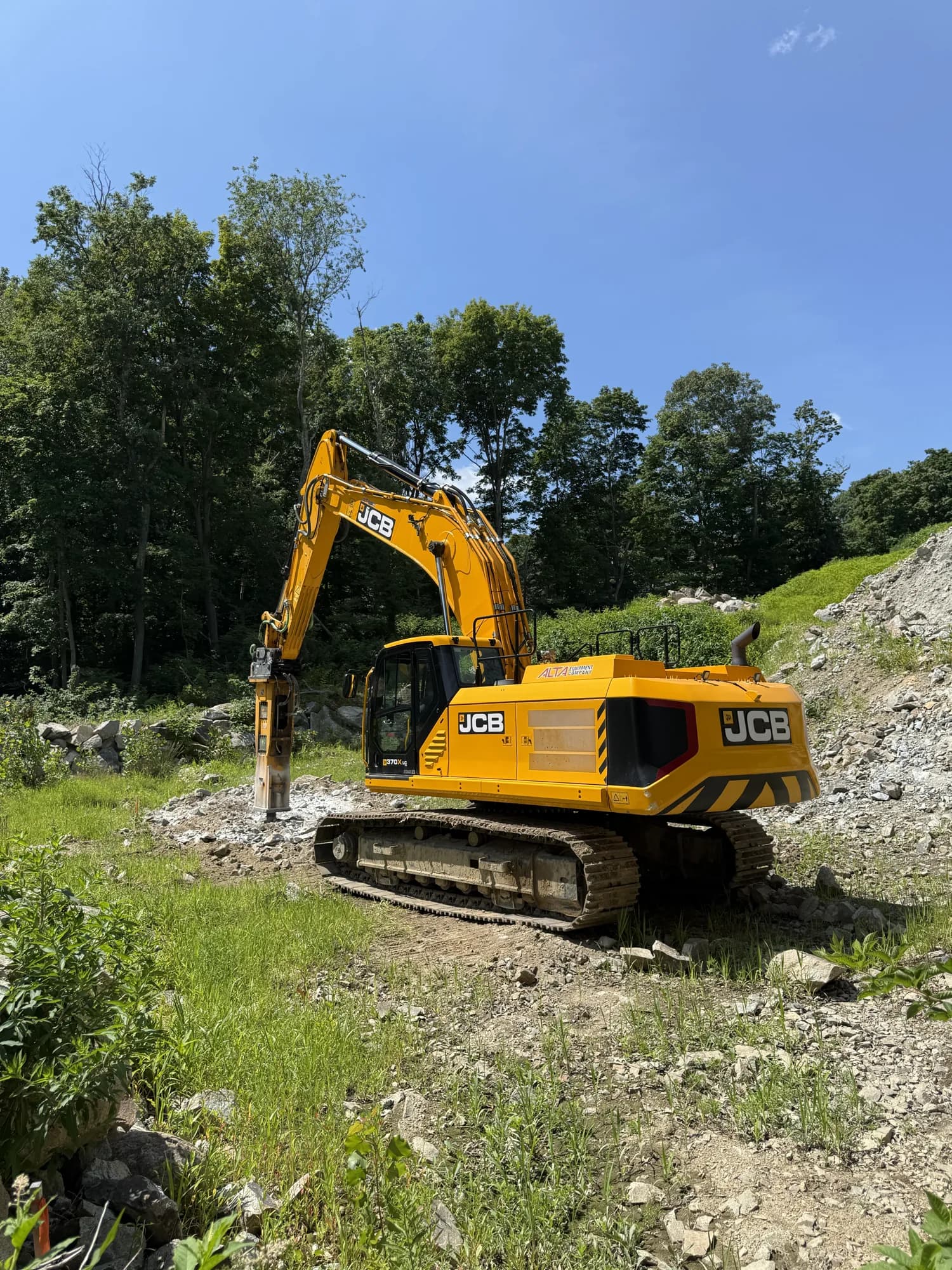 A Coastal Corp JCB tracked excavator running a hydraulic breaker sits at the base of a steep rock embankment on a land-clearing site.