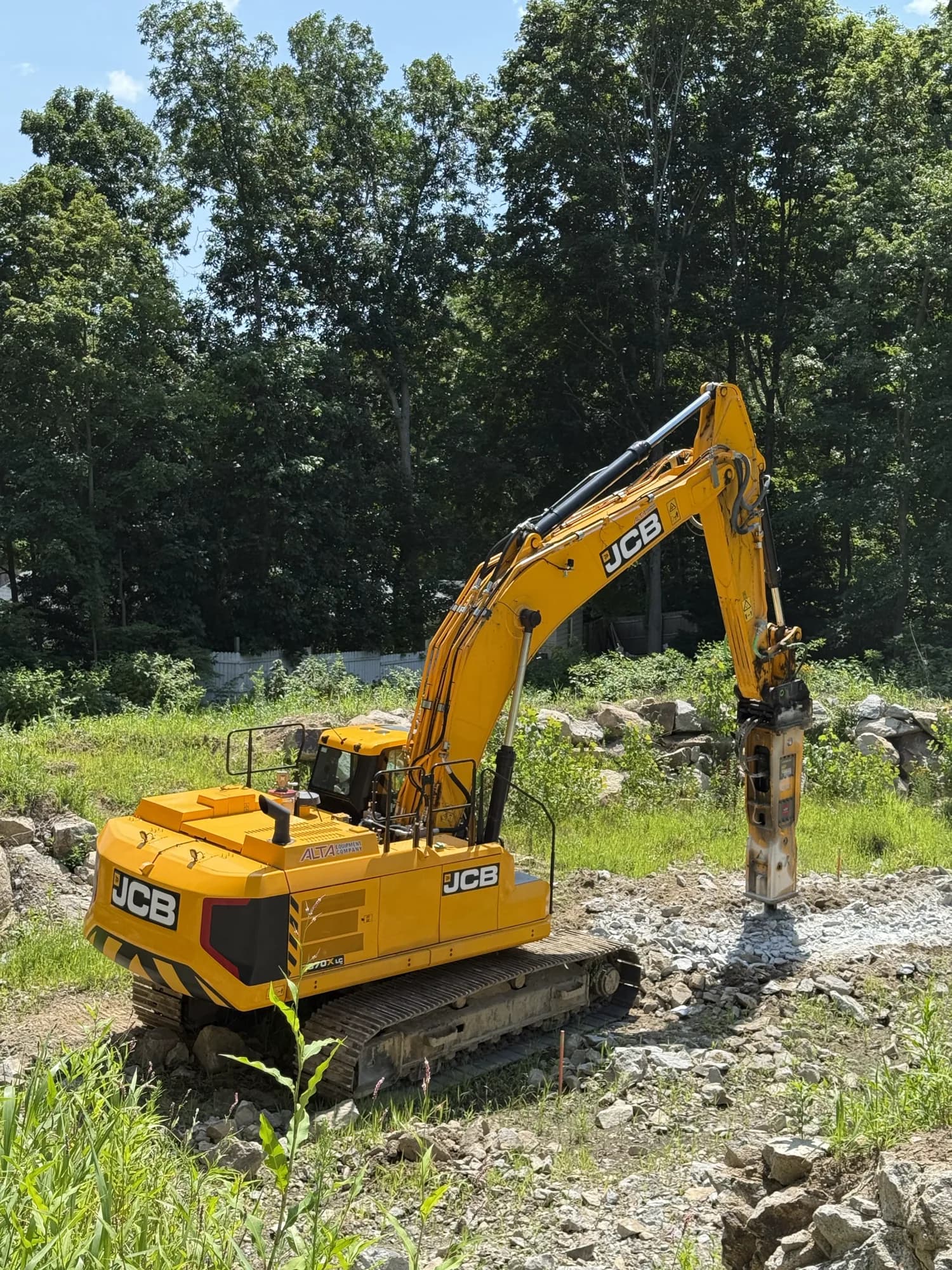 A Coastal Corp JCB tracked excavator fitted with a hydraulic breaker attachment fractures ledge rock on a cleared, wooded jobsite.