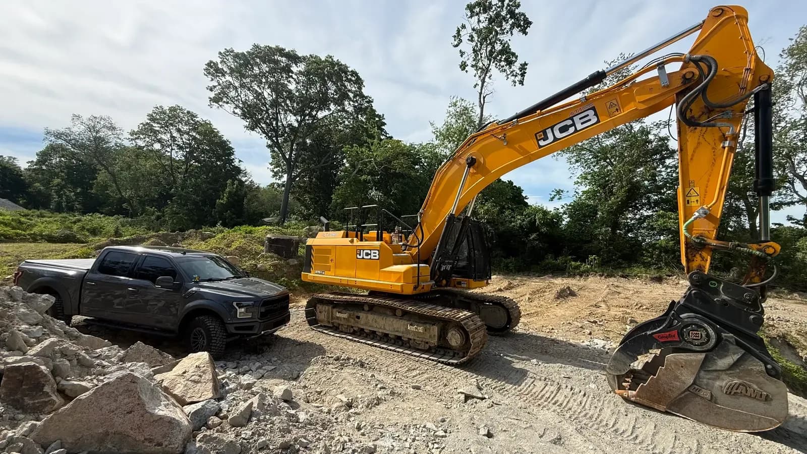 A Coastal Corp JCB tracked excavator with a grapple attachment on a rock and land-clearing site.