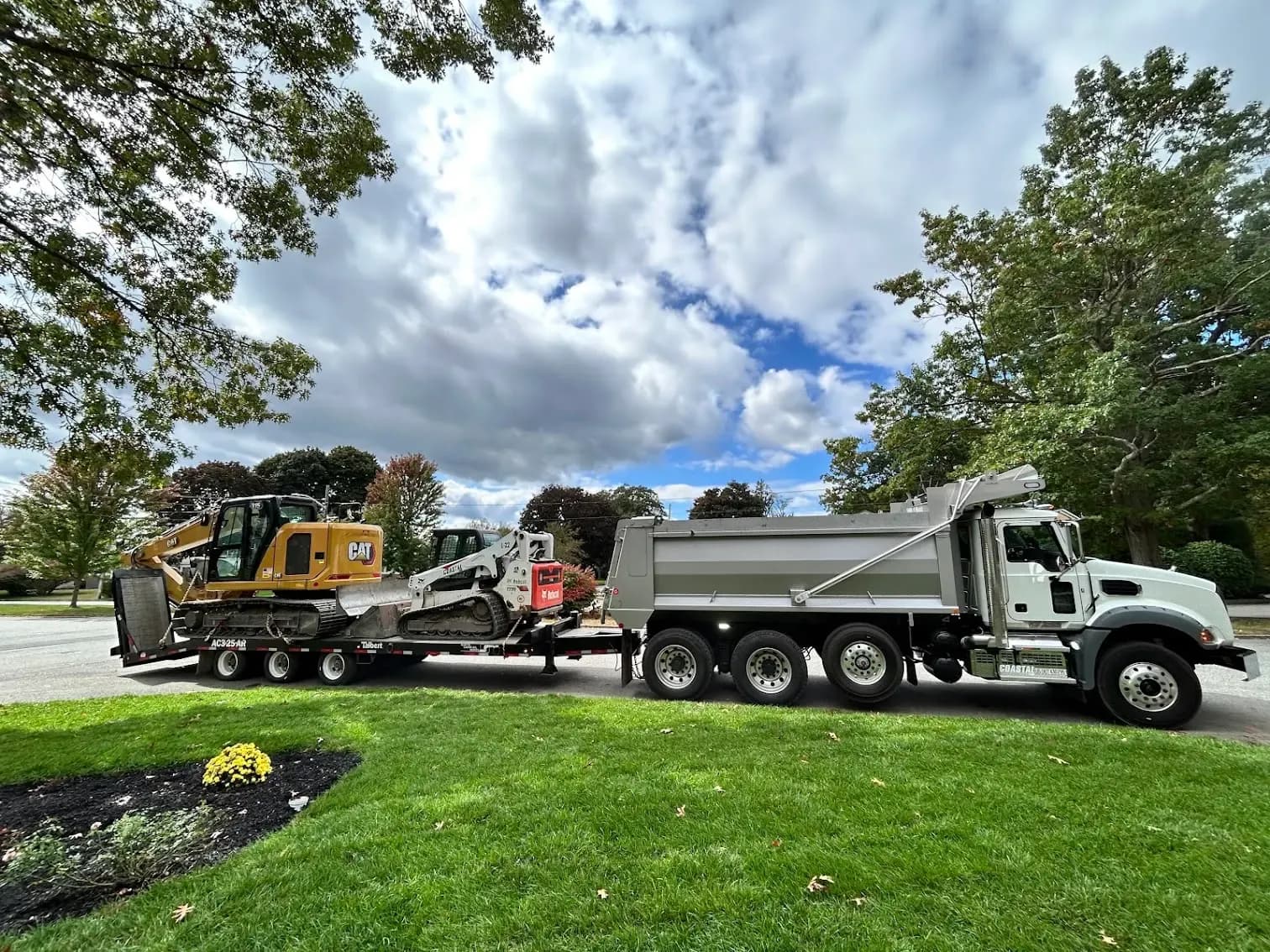 A Coastal Corp Mack tri-axle dump truck hauls a Talbert lowboy trailer loaded with a CAT excavator and a Bobcat track loader.