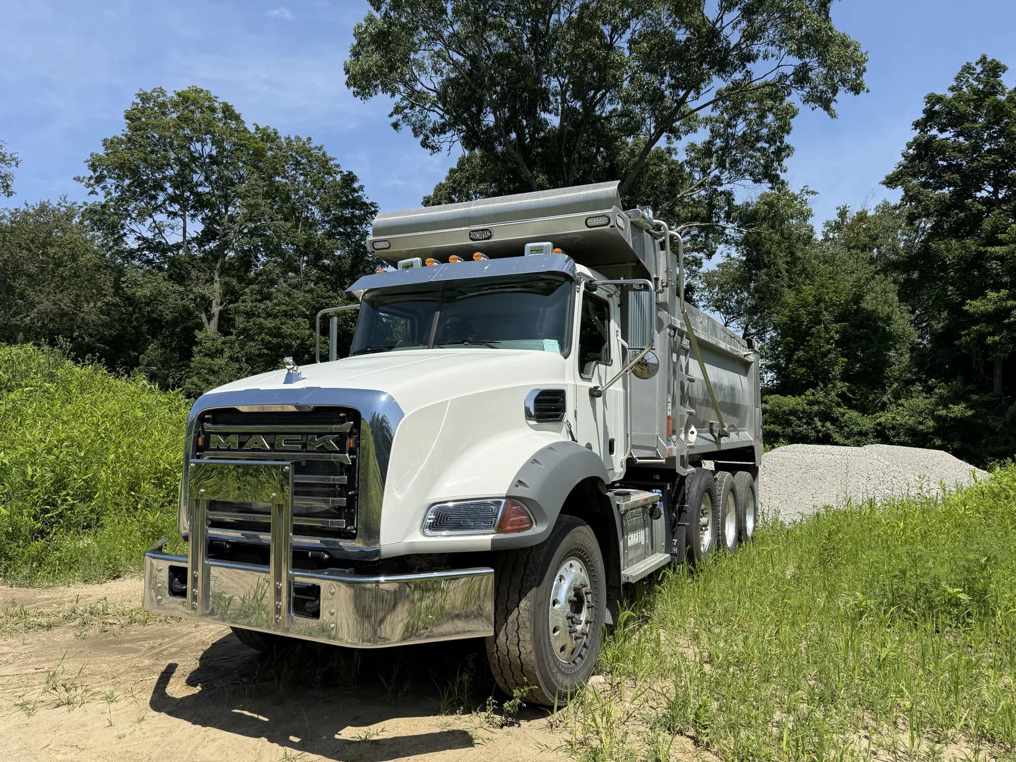 A Coastal Corp white Mack Granite tri-axle dump truck staged on a dirt jobsite beside a fresh gravel stockpile.