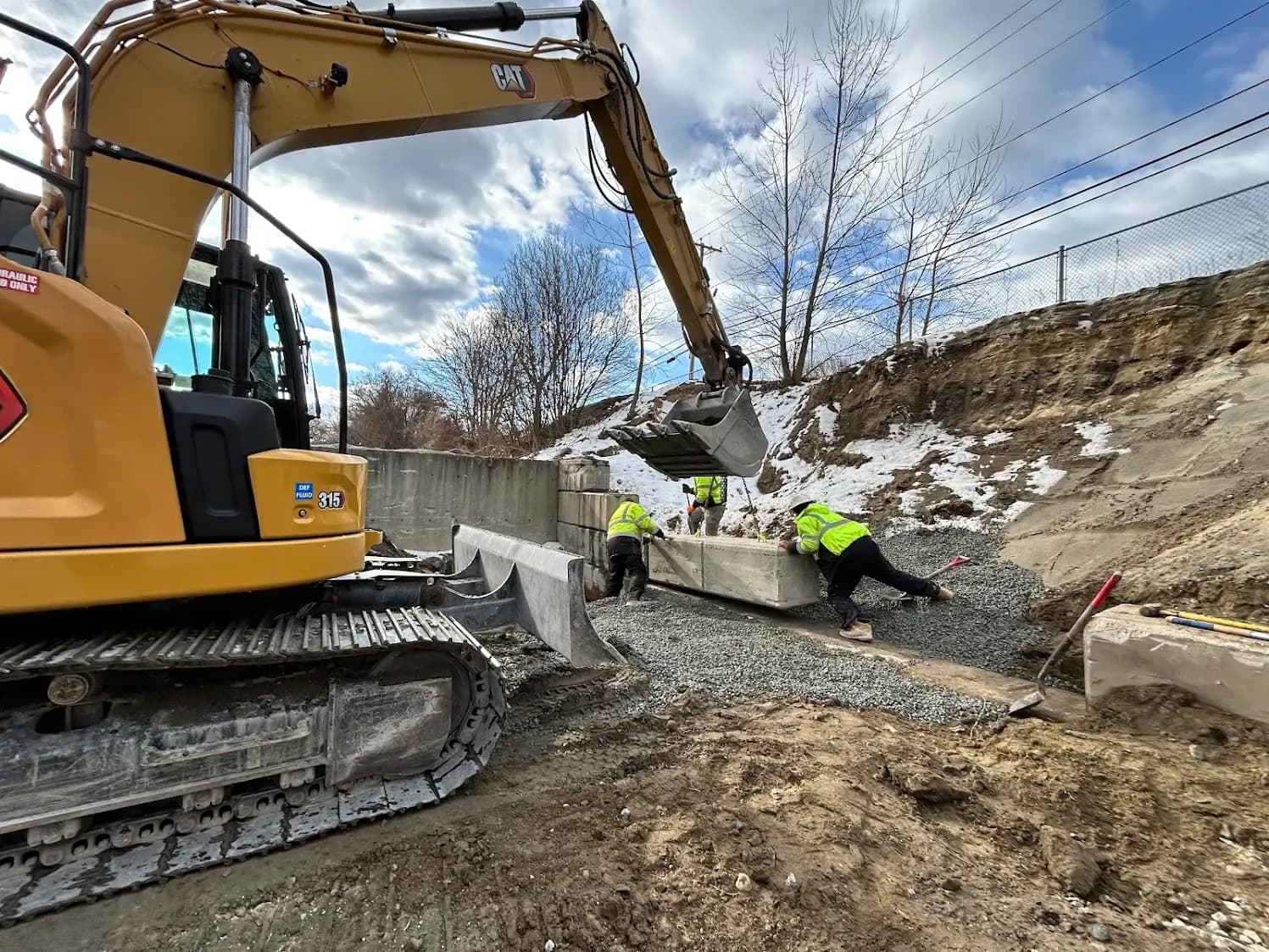 A Coastal Corp CAT excavator sets a precast concrete retaining-wall block while crew members guide it into place on a gravel base.