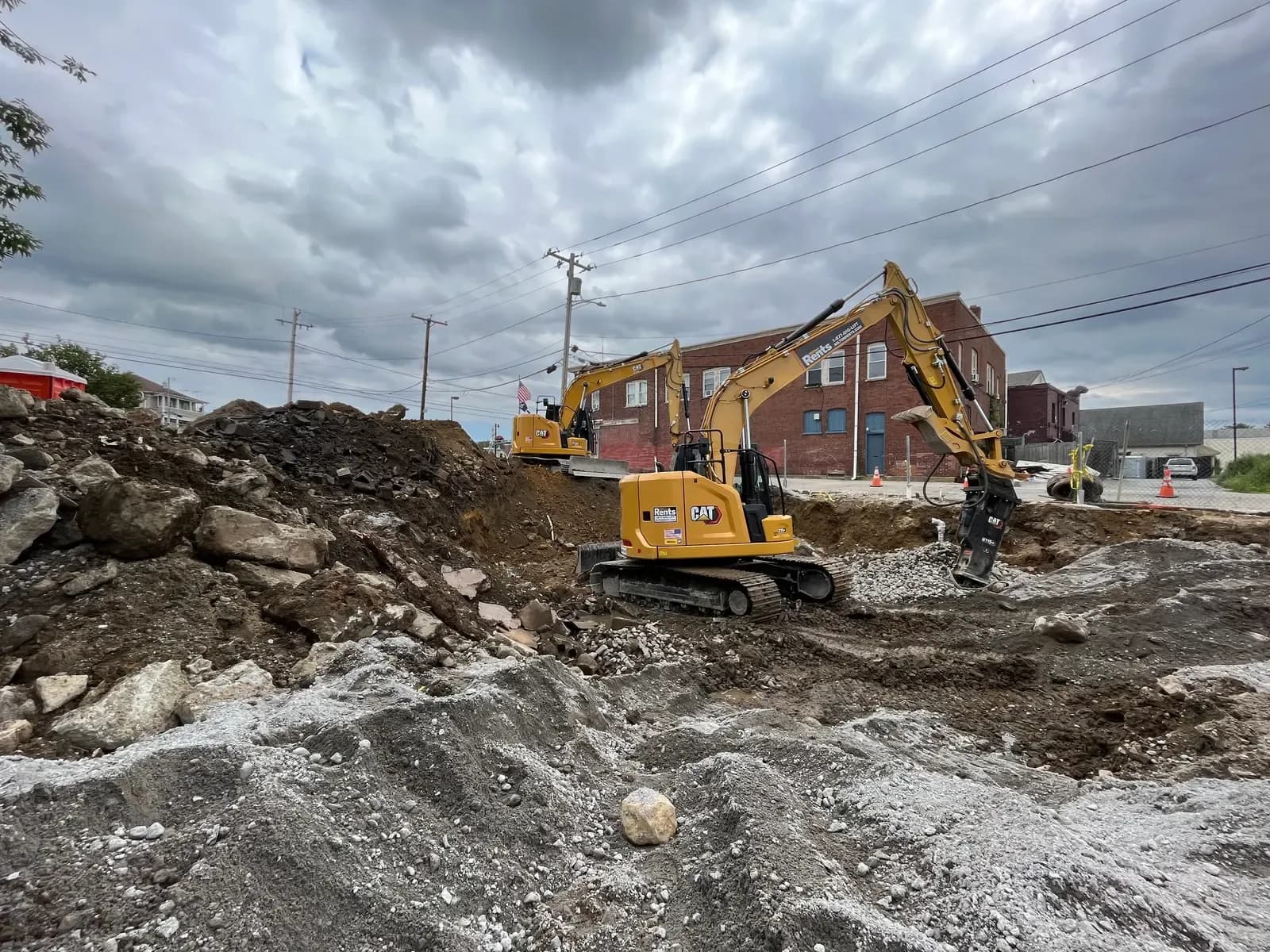 Two CAT excavators work an urban dig, one running a hydraulic breaker to break rock amid piles of broken material.