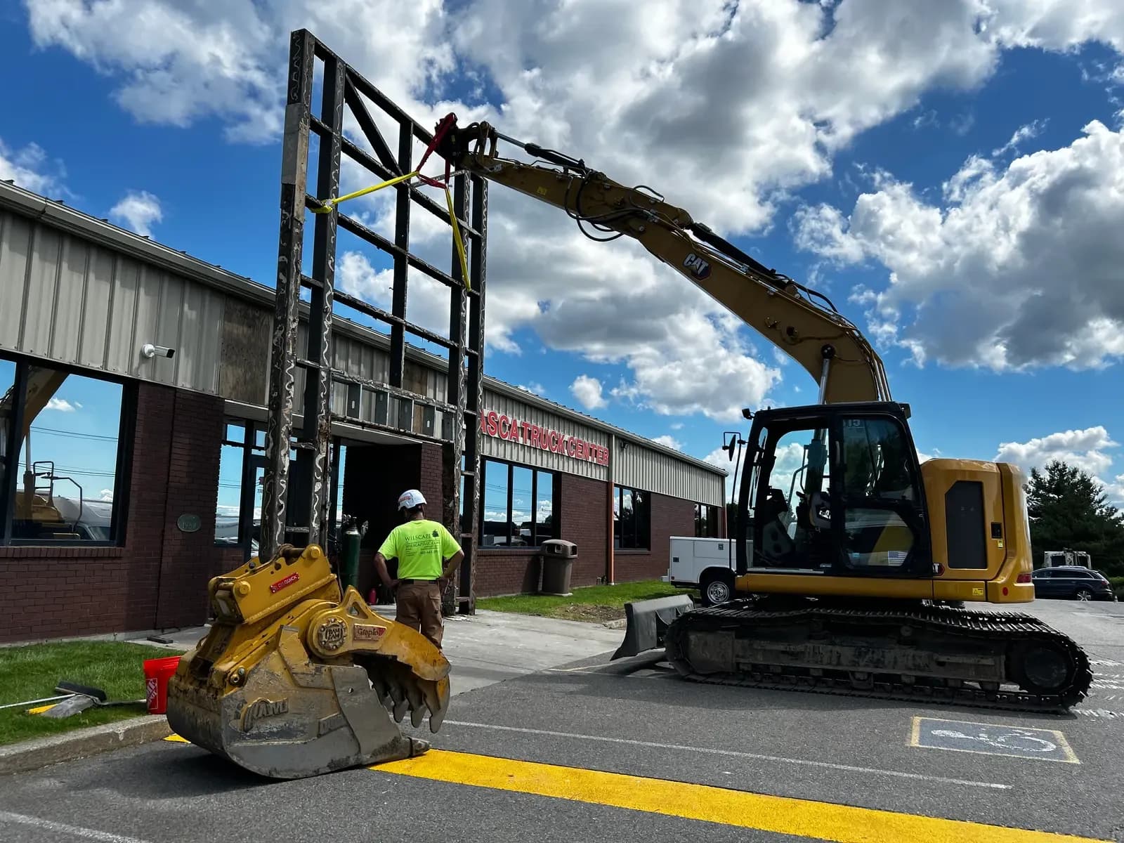 A CAT excavator hoists a large steel sign frame with rigging straps outside the Tasca Truck Center while a worker guides the lift.