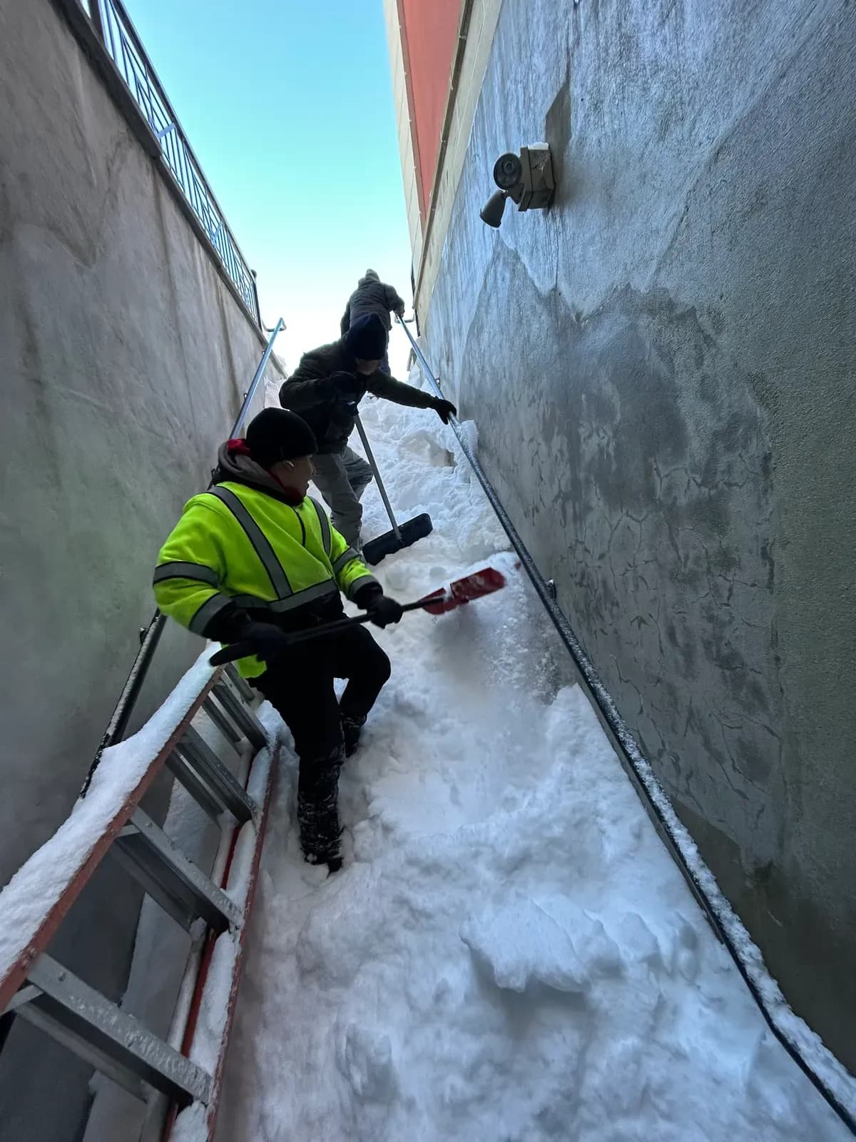 A Coastal Corp crew in hi-vis gear hand-shovels snow from a narrow, tight-access walkway between two commercial buildings.