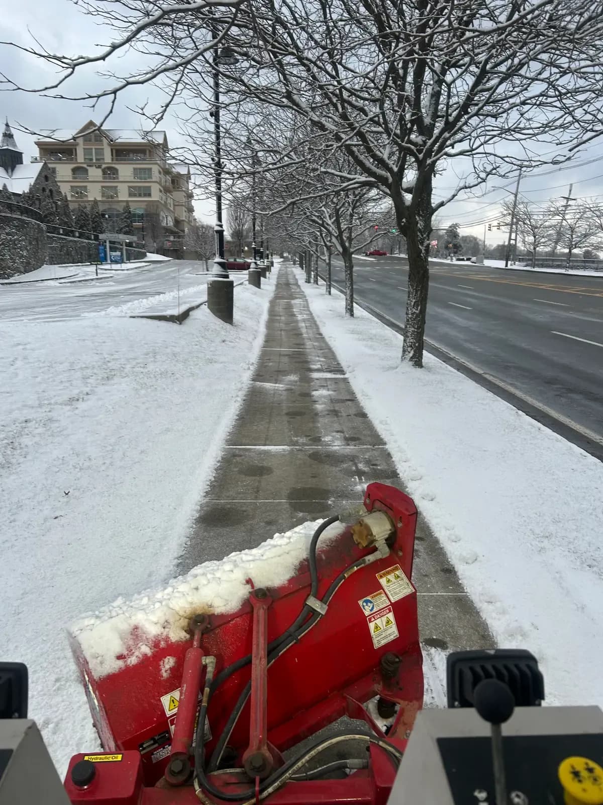 A Coastal Corp sidewalk machine clears a commercial walkway down to bare pavement along a main street lined with trees.