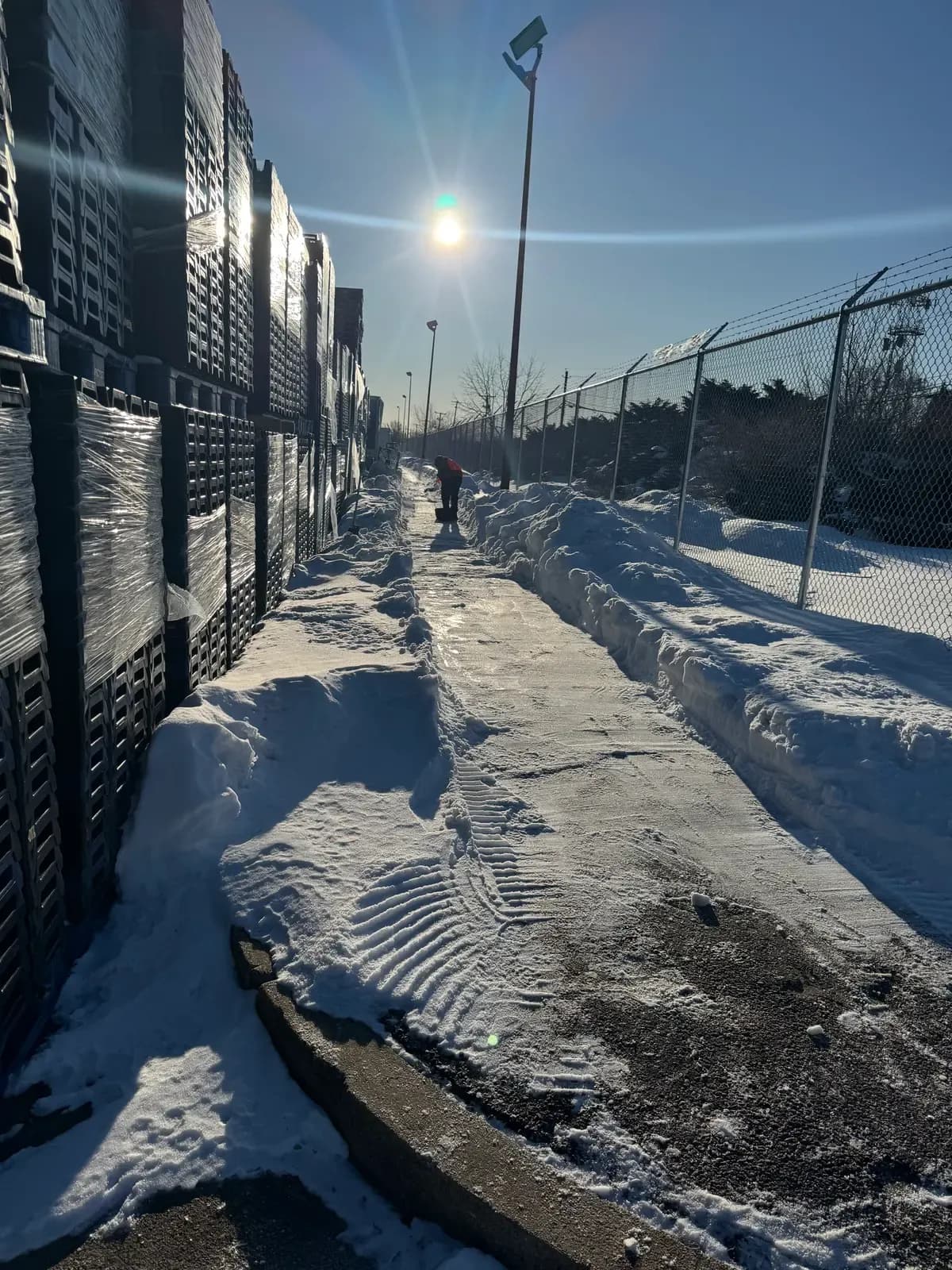 A Coastal Corp worker clears a long industrial-yard walkway along a chain-link fence in bright winter sun after a storm.