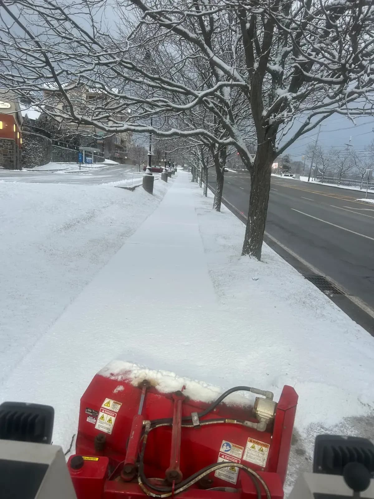 The view from a Coastal Corp sidewalk machine clearing a fresh-snow path along a tree-lined commercial street.
